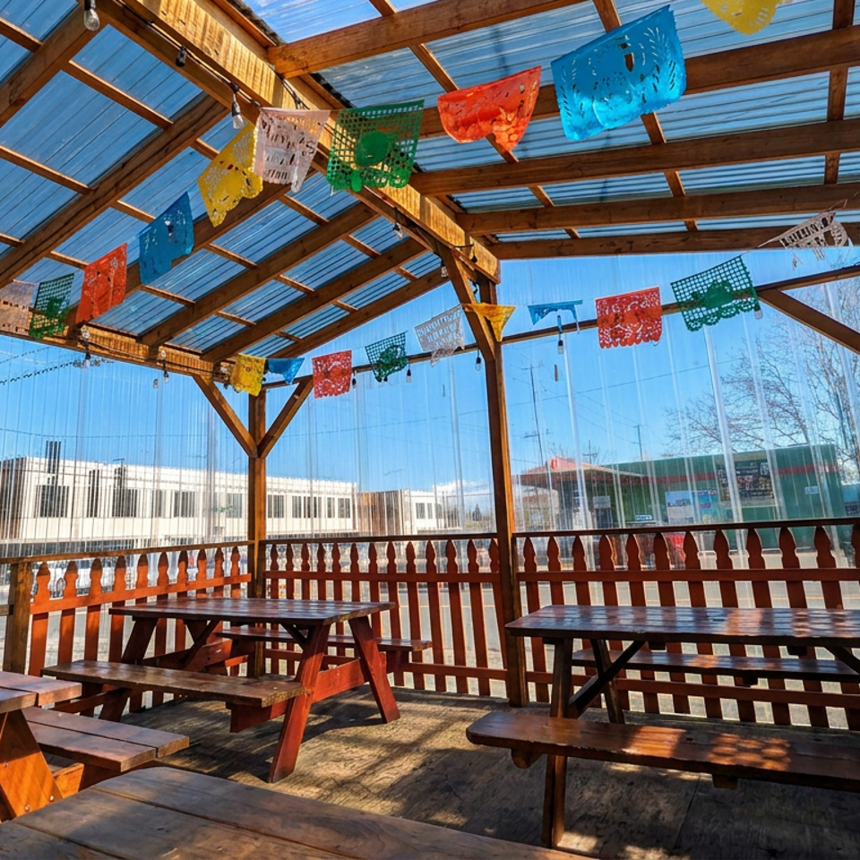 Covered outdoor restaurant patio with wooden picnic tables, colorful papel picado banners, and clear views of surrounding buildings.